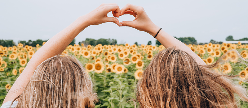 Two girls making a heart by joining their hands together in front of sunflowers